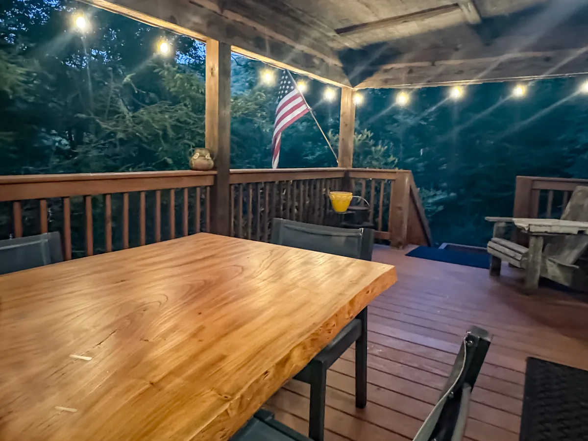 A cozy wooden deck is strung with twinkling lights and features a rustic table and American flag, offering an inviting setting for evening gatherings. The ambiance suggests a seamless transition from day to night, perfect for quiet reflection or celebrating with loved ones beneath the open sky.