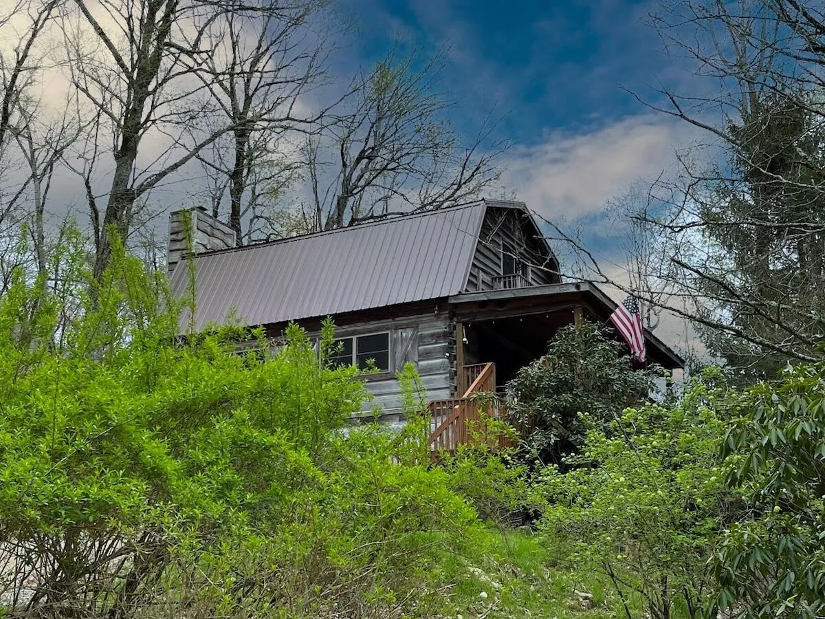 A classic log cabin stands nestled among lush greenery, its inviting porch adorned with an American flag. The surrounding woods create a peaceful, private setting that hints at the adventures waiting just beyond the cabin's doorstep.