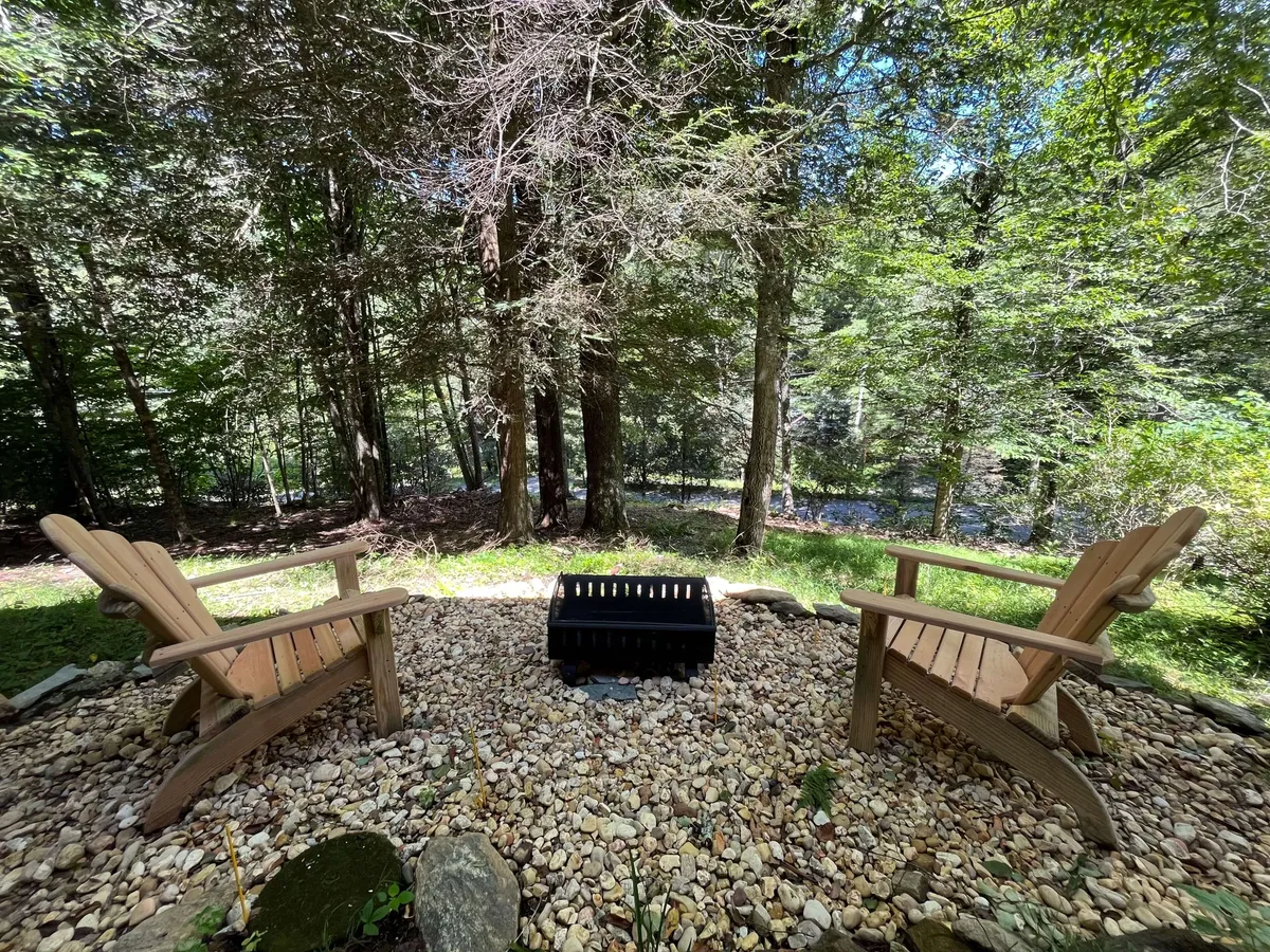 Two wooden chairs circle a rustic fire pit set amongst tall trees, with pebbled ground underfoot. The serene outdoor setup invites relaxation, conversation, and stargazing, surrounded by nature.