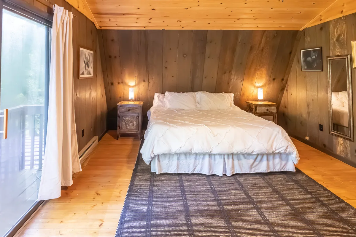 A light-filled bedroom featuring a plush bed with white bedding, nightstands, a large window that frames tranquil forest scenery, a cozy rug, and simple wall art. The room feels intimate and soothing, ideal for quiet relaxation.