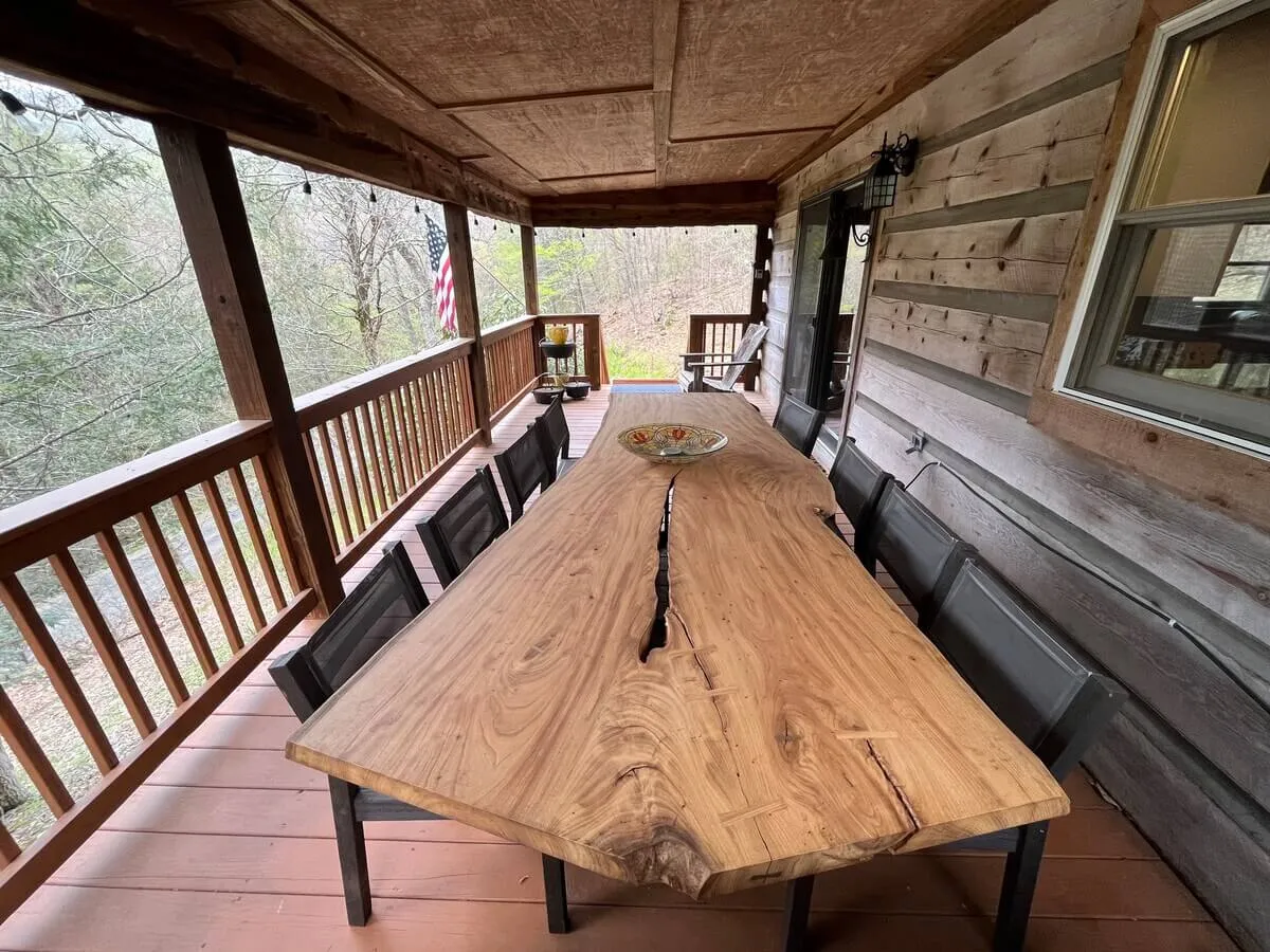 A large wooden table sits on a deck overlooking lush greenery, with a sturdy wooden railing that encloses the space. The setting is perfect for outdoor dining, morning coffee, or evening drinks in the fresh air, with ample space for gatherings or quiet moments.