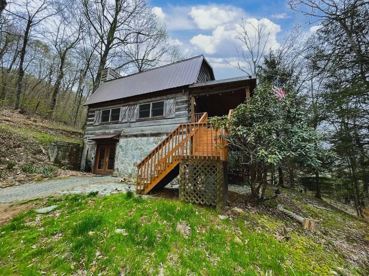 A welcoming exterior shot of the log cabin showcases its stone and wood facade surrounded by trees, with a wooden staircase leading up to a cozy porch. The peaceful setting highlights the cabin's privacy and immersion in nature.