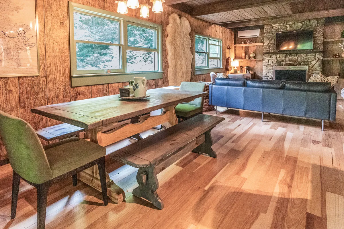 A warm dining area featuring a sturdy wooden table set near a stone fireplace, complemented by a blue leather sofa and large windows that flood the space with natural light. The ambiance is inviting and perfect for shared meals or evenings of games and laughter.