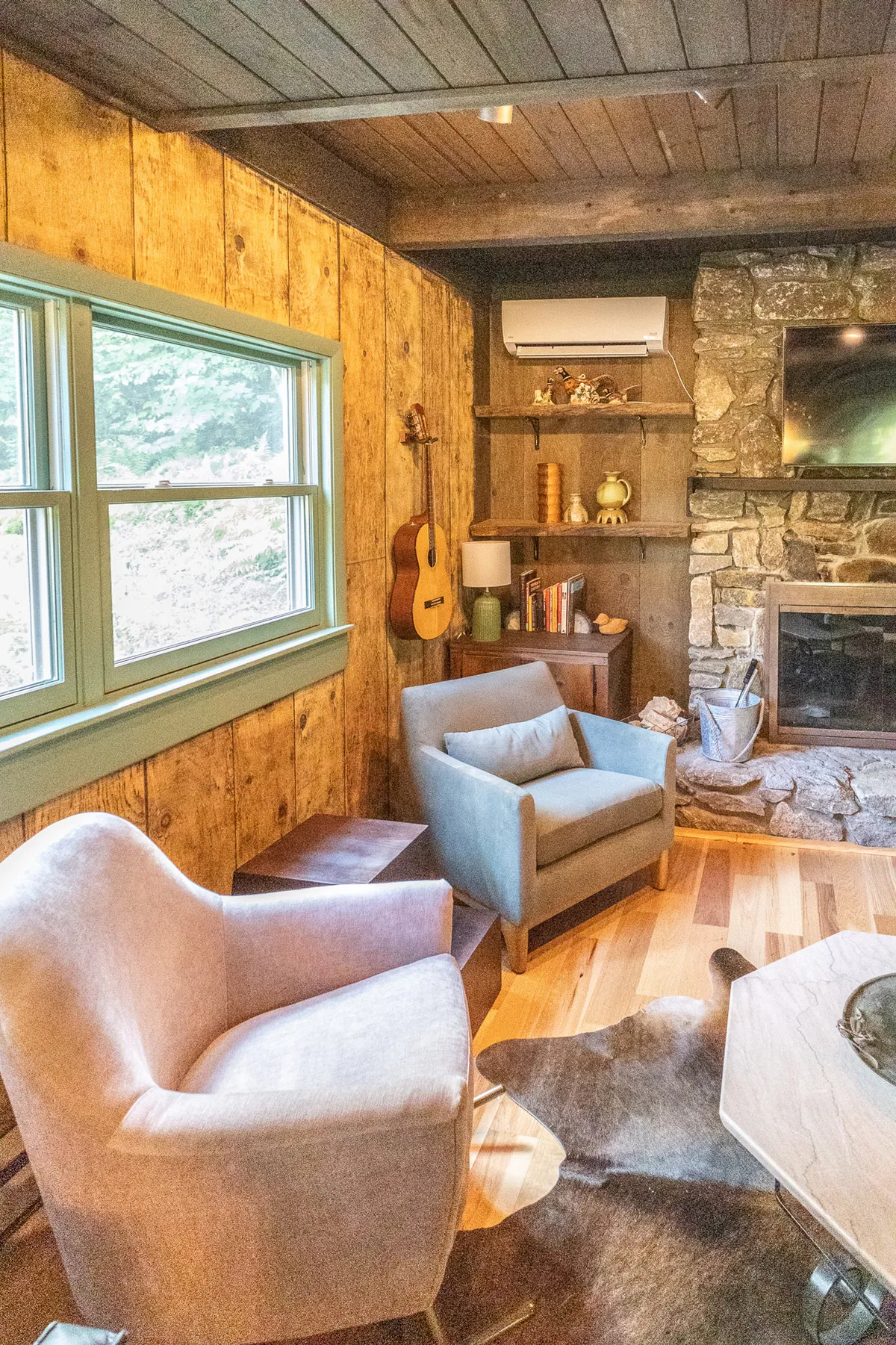 A welcoming living area featuring wooden walls, a stone fireplace, a guitar resting nearby, shelves adorned with decorative items, and plush, comfortable seating. Large windows bathe the room in natural light and provide glimpses of the surrounding forest.