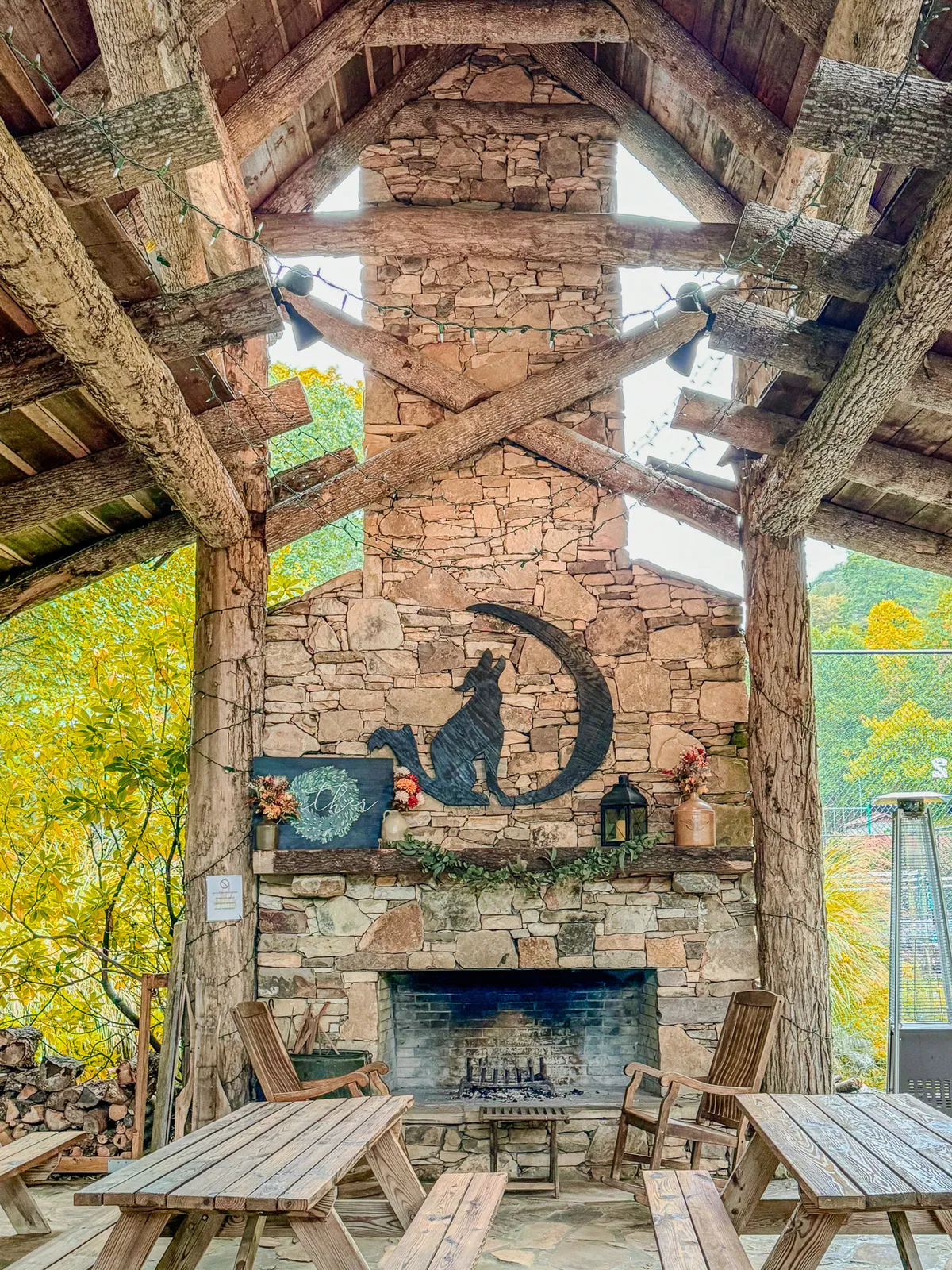 This image showcases a rustic living room with exposed log beams, a prominent stone fireplace at the center, and thoughtfully chosen decor including a cat silhouette artwork and sturdy wooden tables. The space is filled with natural light and exudes warmth, with inviting seating arrangements perfect for gatherings.