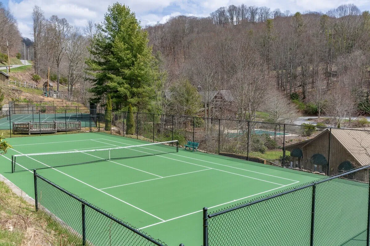 An outdoor tennis court surrounded by tall, leafy trees, with benches for spectators or resting players. The court is clean and ready for friendly matches, capturing the energy of outdoor play.