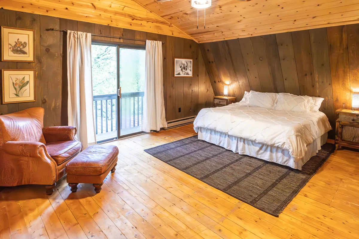 A tranquil bedroom with a comfortable bed positioned near a sliding glass door, a leather chair for relaxing, and tasteful artwork on the walls. Soft natural light filters through the glass, illuminating gentle wood tones and inviting textures, with greenery from the outdoors visible just beyond.