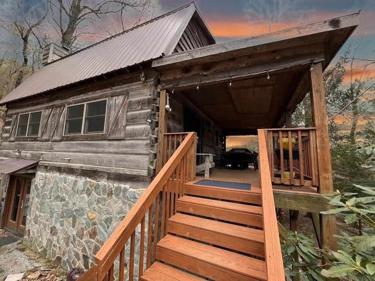 The hero image features a charming antique log cabin set among towering trees, with a classic wooden porch, sturdy stone foundation, and inviting steps leading up to the entry. The natural surroundings evoke a feeling of seclusion and tranquility, highlighting the cabin’s unique rustic character and mountain setting.