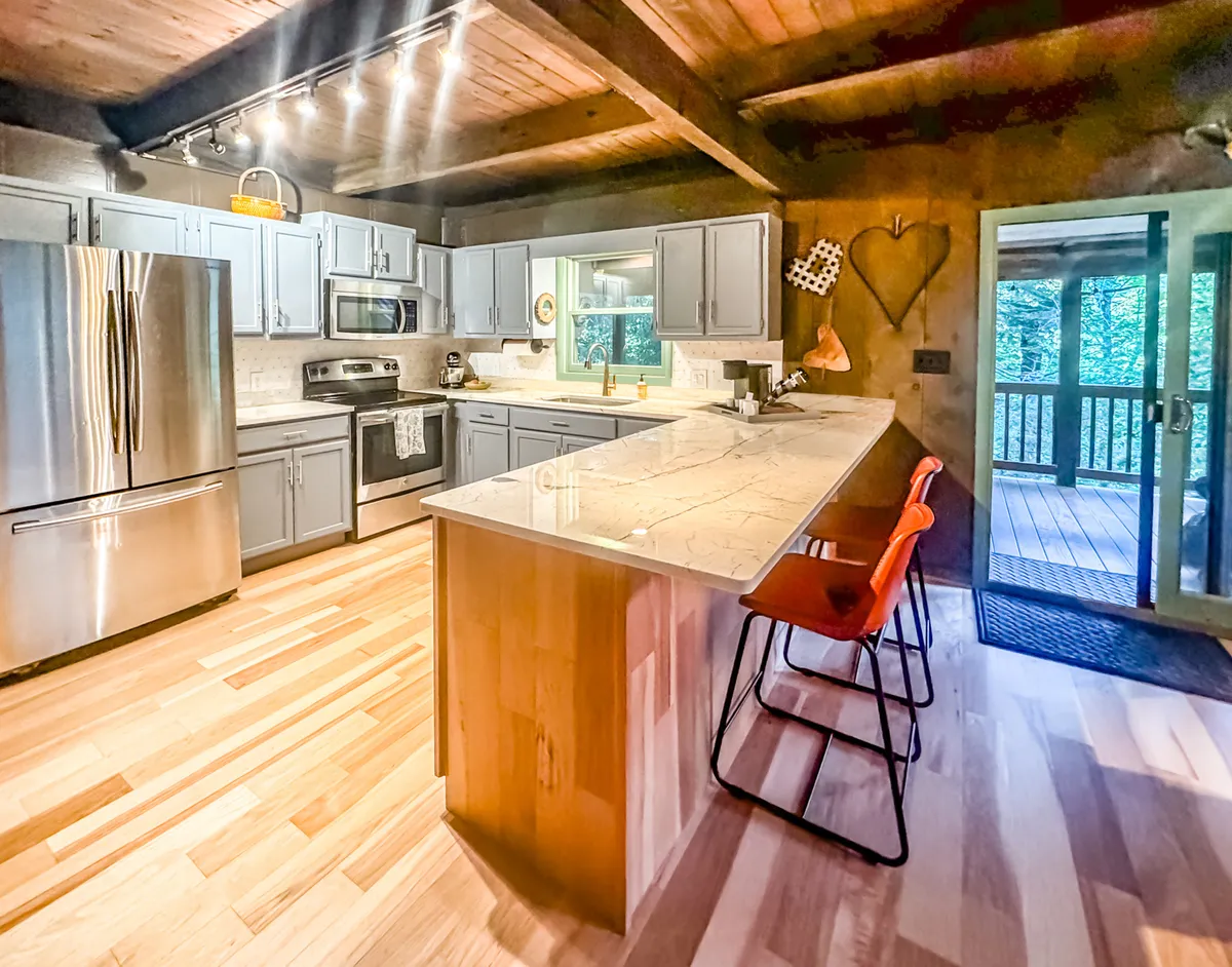 A cozy, updated kitchen with an island countertop, gleaming stainless steel appliances, and rustic wooden beams overhead. The layout is open, with space for cooking and entertaining, and subtle details hint at the cabin’s original charm.